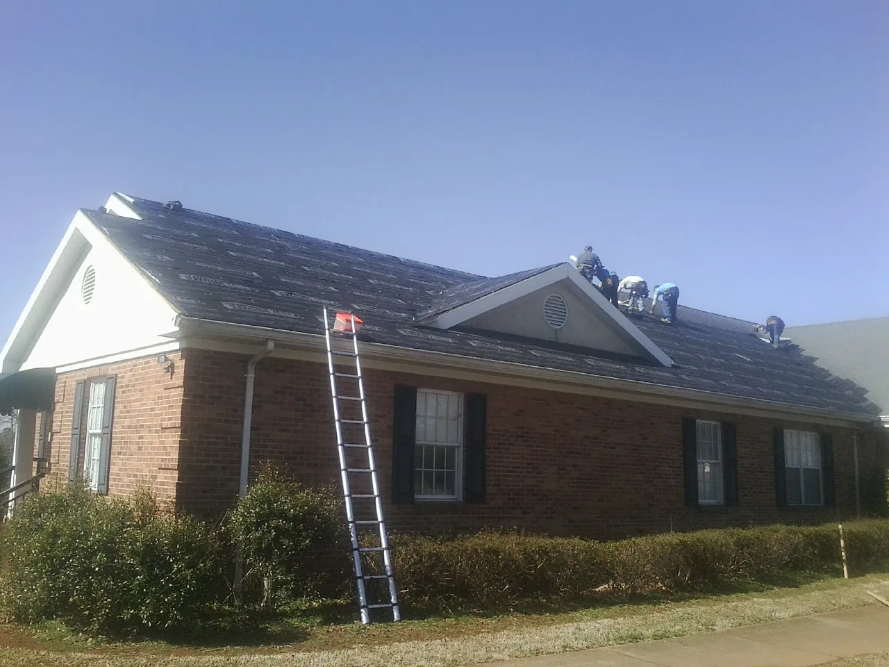 Crew tearing shingles off a brick ranch roof with a ladder set — Emergency Roof Repair work by our crew in Maybee