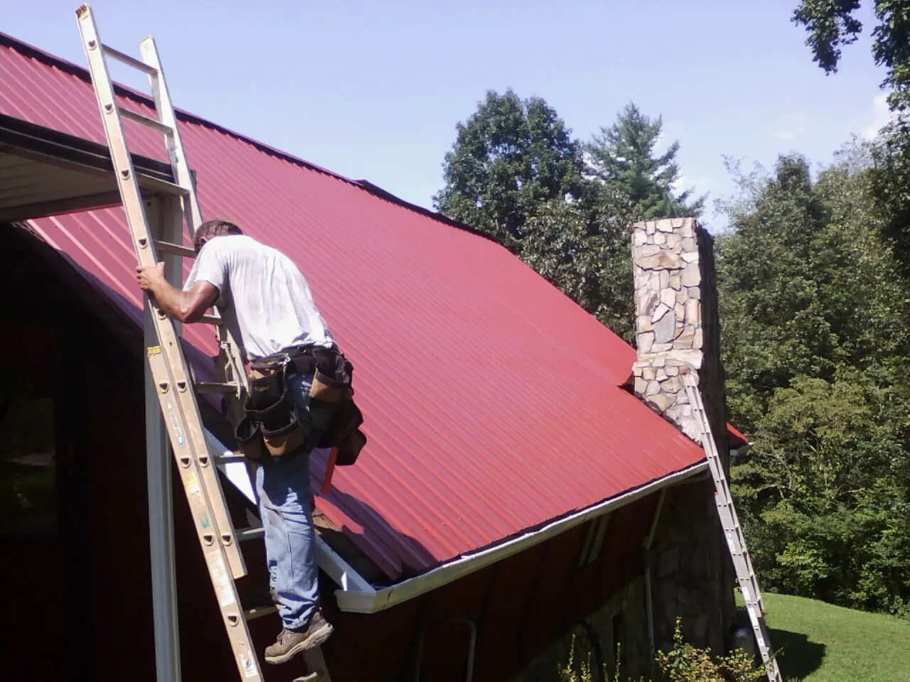 Roofer on a ladder beside a red metal roof and stone chimney — Soffit & Fascia Repair by Roofing Brothers in Maybee